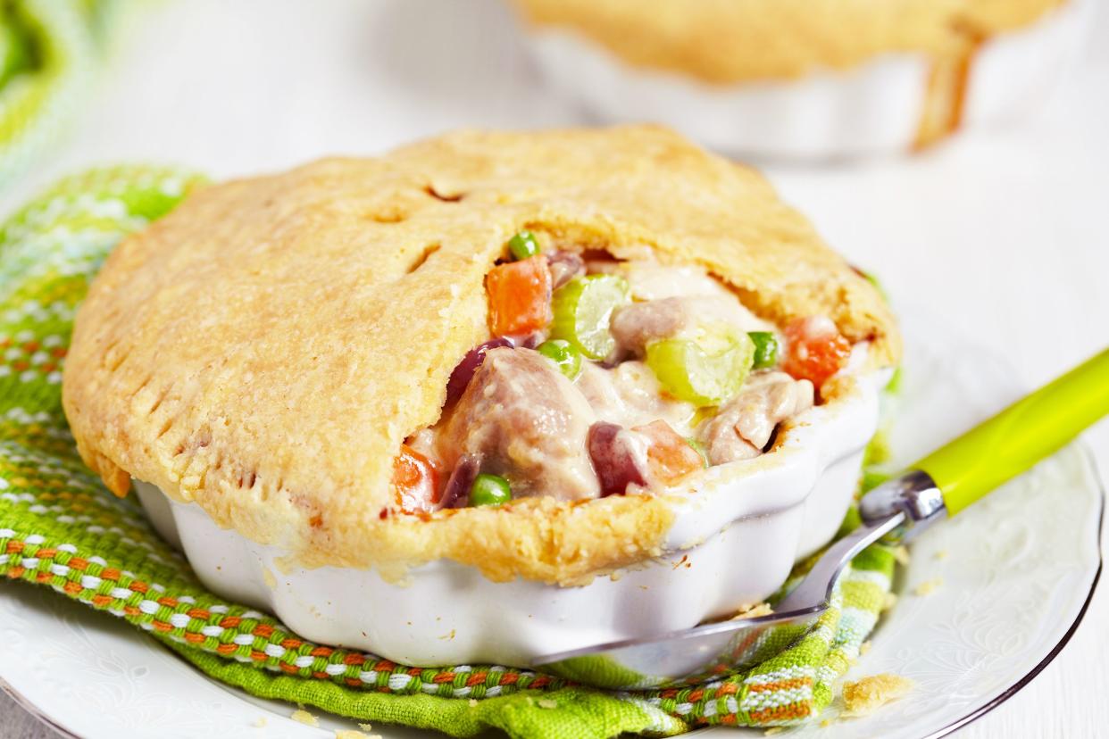 Chicken pot pie in a small white porcelain dish on a avocado green napkin with a matching spoon on a gold rimmed porcelain plate with another one in the background