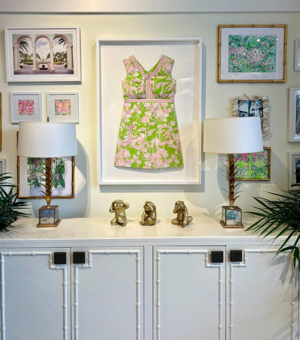 a white kitchen dresser with lamps on it next to a wall with a framed dress and framed colorful prints