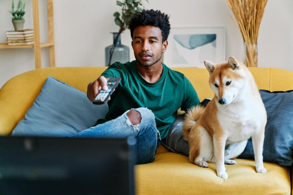 Man sitting on sofa in living room with dog watching TV changing channels with remote control.