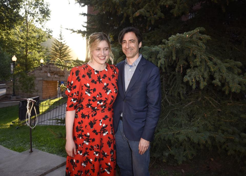 greta gerwig and noah baumbach pose for a photo together while standing in front of an evergreen tree, both smile and gerwig looks right of the camera, she wears a red dress with a black and white floral pattern, he wears a navy suit jacket, gray collared shirt and navy pants