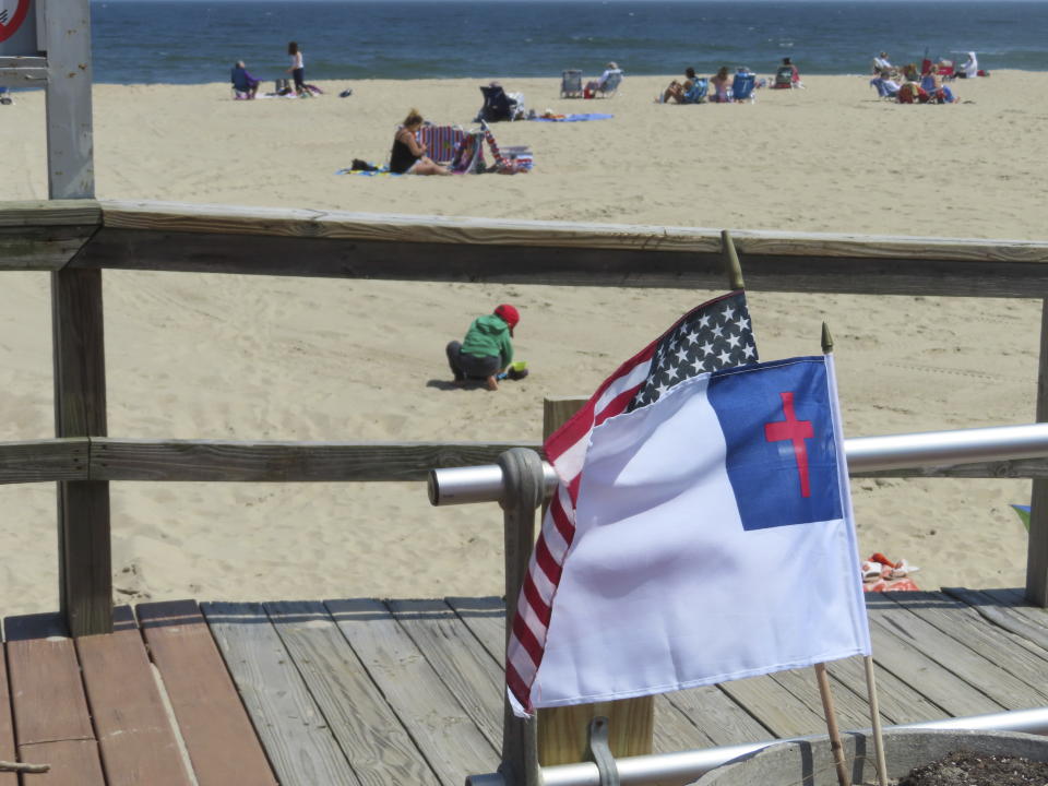 People sit on the beach on a weekday afternoon in Ocean Grove, N.J. on May 2, 2024 as a Christian flag and an American flag flutter in the breeze. The state of New Jersey says the Ocean Grove Camp Meeting Association is violating state beach access laws by keeping people off the beach until noon on Sundays. (AP Photo/Wayne Parry)