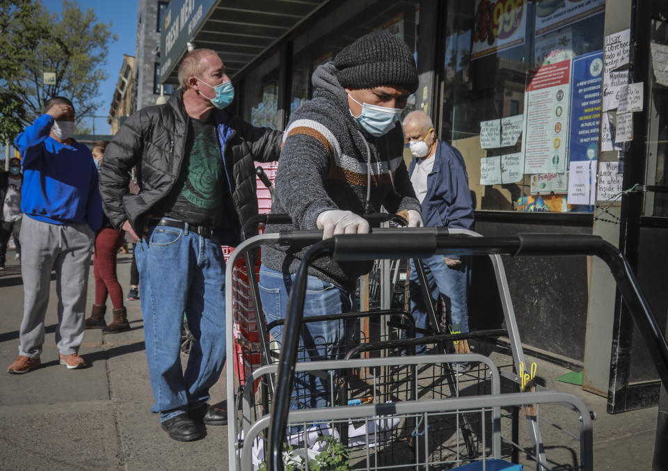 A masked grocery store worker in Brooklyn's Sunset Park—a neighborhood with one of the city's largest Mexican and Hispanic community, organize shopping carts while people waiting to enter the store also wear masks to help stop the spread of coronavirus, Tuesday May 5, 2020, in New York. A poll found that 61% of Hispanic Americans say they've experienced some kind of household income loss as a result of the COVID-19 outbreak. (AP Photo/Bebeto Matthews)