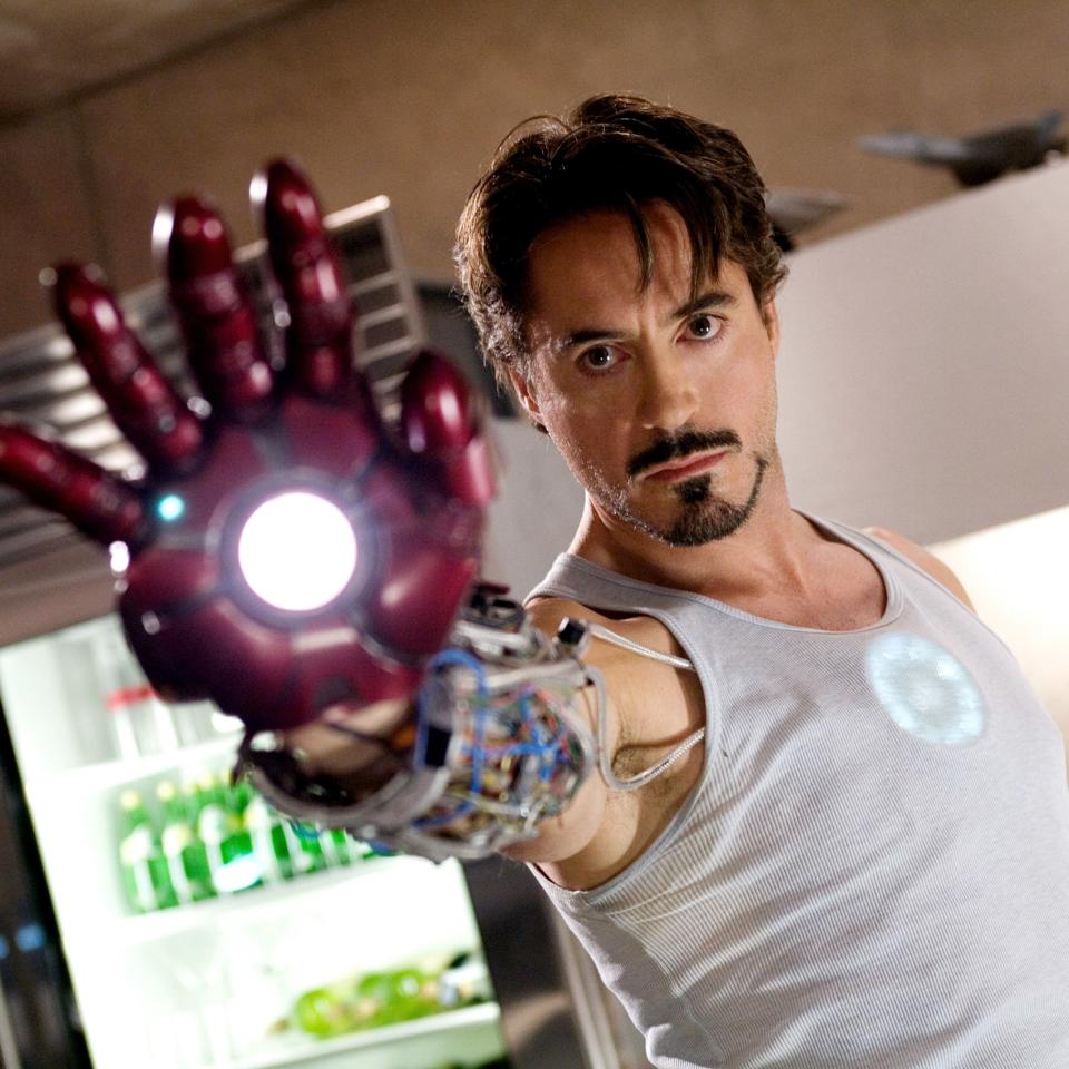 Robert Downey Jr. as Tony Stark/Iron Man points his arm with a glowing hand repulsor forward, wearing a tank top in what appears to be a lab or high-tech setting