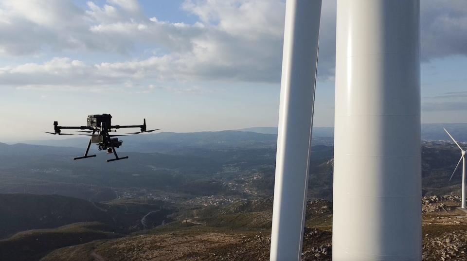 A black drone flies near a white wind turbine in the mountains.