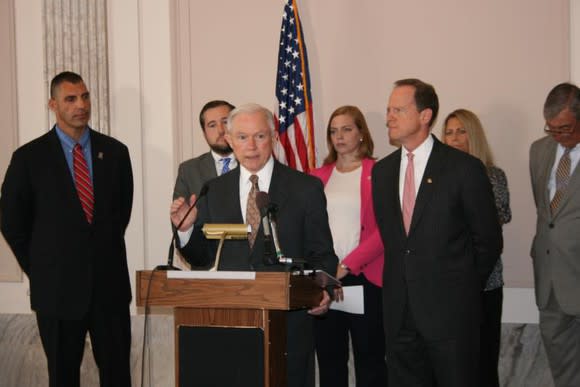 Jeff Sessions addressing an audience from behind a lectern with six people standing behind him.