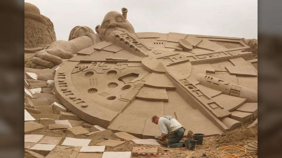 A man sculpts sand into an enormous Millennium Falcon with Shrek in the background.