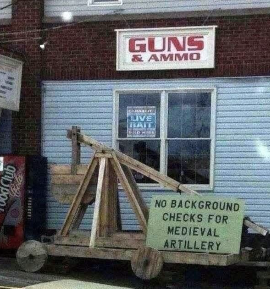 "No background checks for medieval artillery" sign on an ancient military device for hurling missiles outside a "Guns & Ammo" store