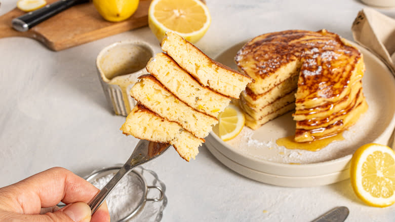 Holding a fork with a stack of pancakes over a plate with pancakes and lemons on the side