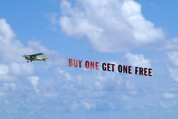 Airplane is towing a banner that reads 'Buy One Get One Free' against a beautiful sky with clouds.