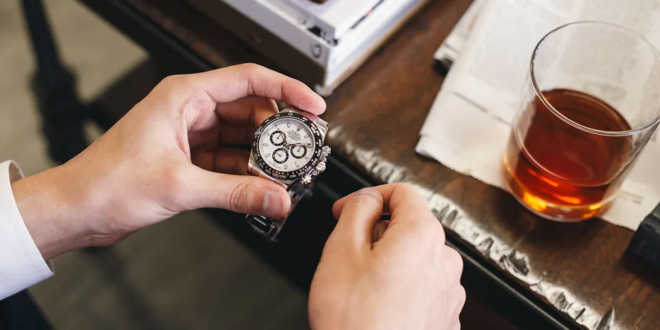 A man holding a Rolex Daytona watch with a glass of whiskey on the table.