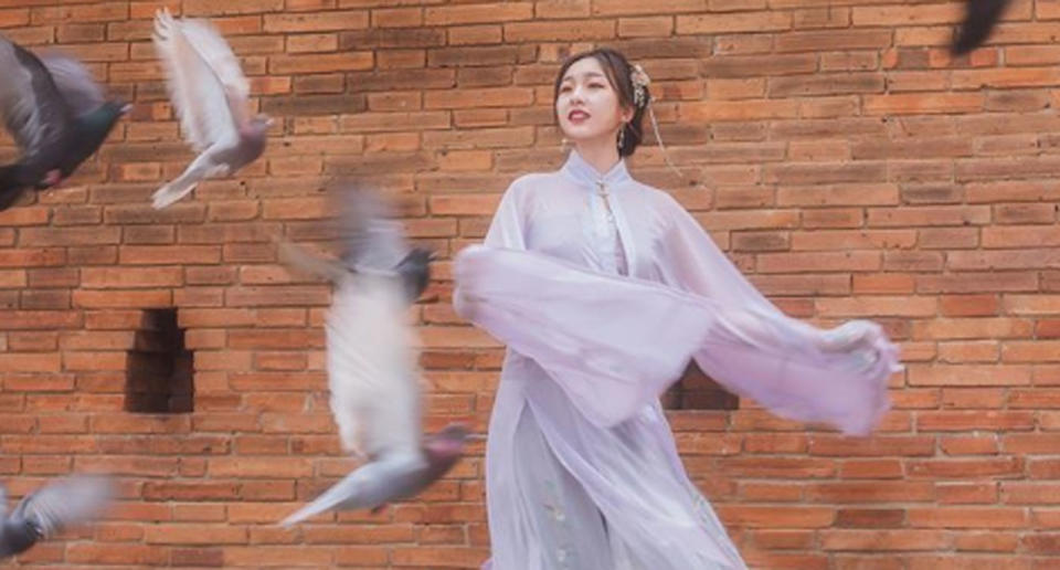 A woman stands at Chiang Mai's Tha Pae Gate in Thailand posing for a photo as pigeons fly past.