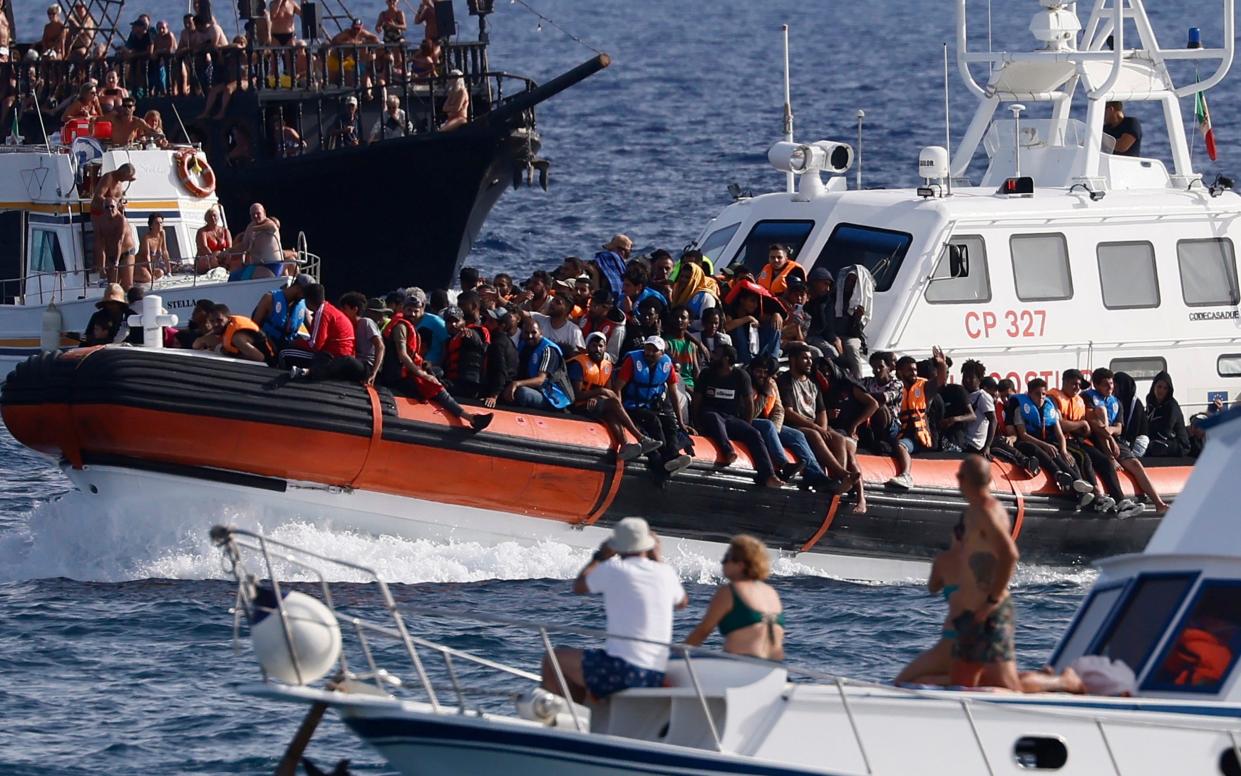 An Italian Coast Guard boat carries migrants as tourists on boat, foreground, watch, near the port of the Sicilian island of Lampedusa,