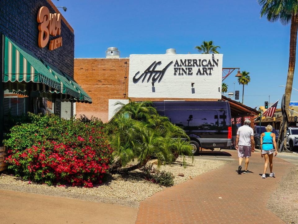 A brick path with storefronts and bushes on the left and a tree on the right on a clear day with blue skies