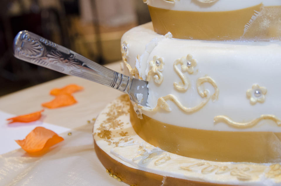 Close-up of a decorated wedding cake being cut with a silver knife, showcasing intricate fondant designs and orange flower petals on the table