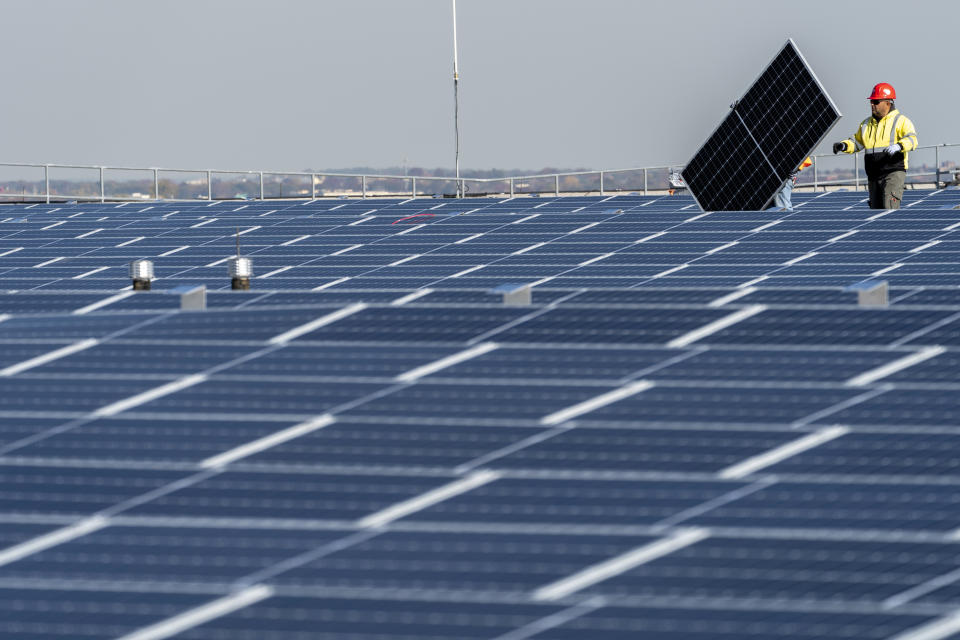 A worker in a red hardhat walks across a solar array followed by a colleague carrying a solar panel.