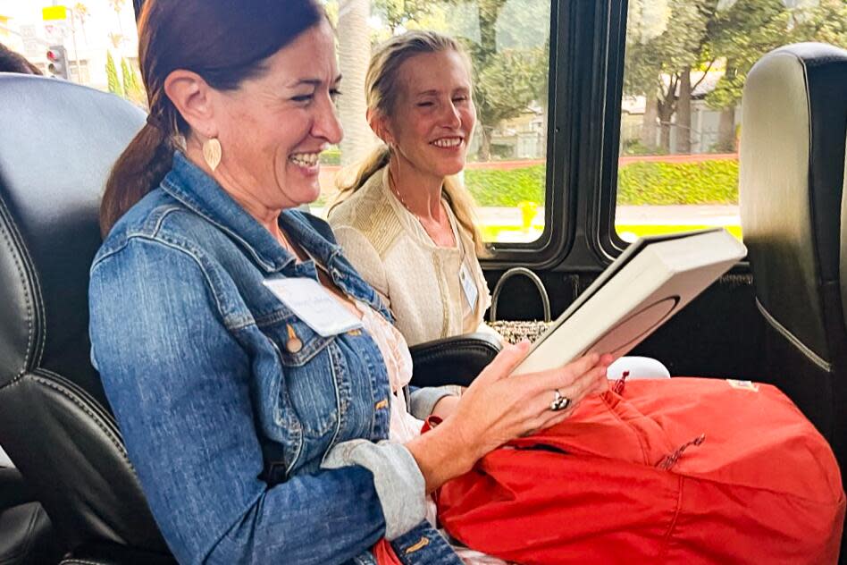 Two smiling women sit side by side on a bus, one holding a book