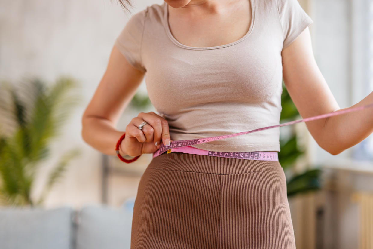 Beautiful young woman measuring her waist with a measuring tape in the living room at home. BMI