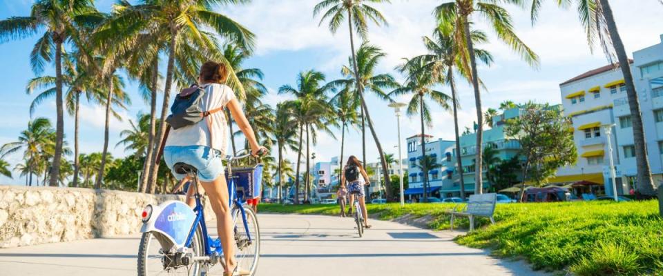 MIAMI - CIRCA JUNE 2017: Young woman rides bike along the Miami Beach promenade with backdrop of iconic art-deco buildings and palm trees of Ocean Drive