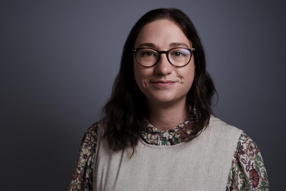 A white woman with glasses and dark, medium-length wavy hair wears a floral top and smiles slightly at the camera.