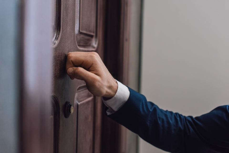 Person in a suit knocking on a wooden door. The hand is visible while the rest of the body is not fully shown