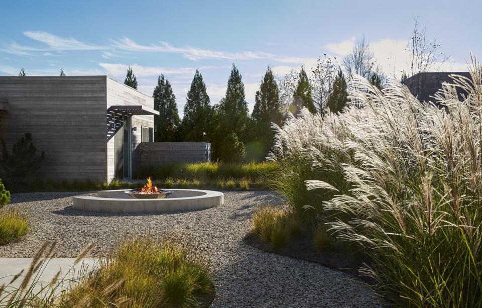 garden area with circular patch covered in gravel and a center fountain and wispy plants on the right and a small square gray house on the right background