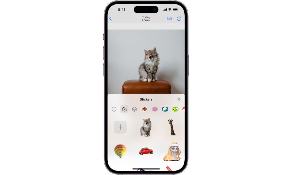 A screenshot inside an iPhone frame, showing the Stickers pop-up menu in the iOS Photos app. A cat yawns in the photo, and the stickers drawer below shows it among others (including a giraffe, balloon, car, etc.).