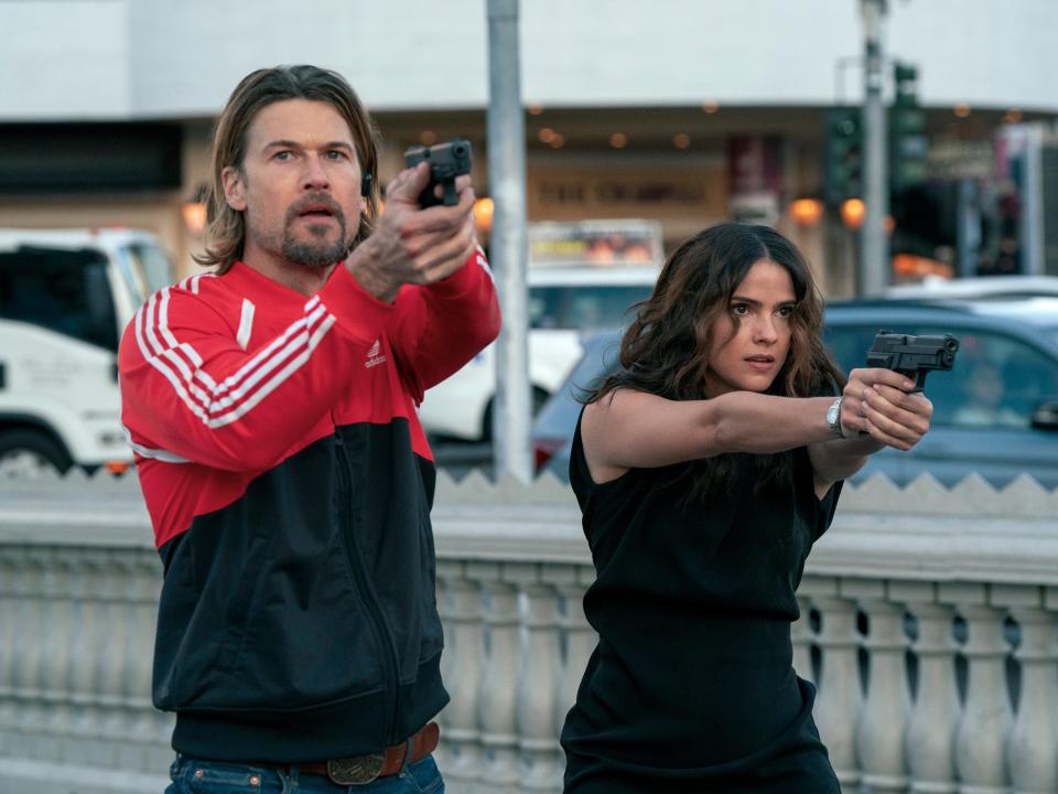 nick zano as chad mcknight and shelley hennig as ava winters in obliterated, both intently aiming their guns while standing near a road