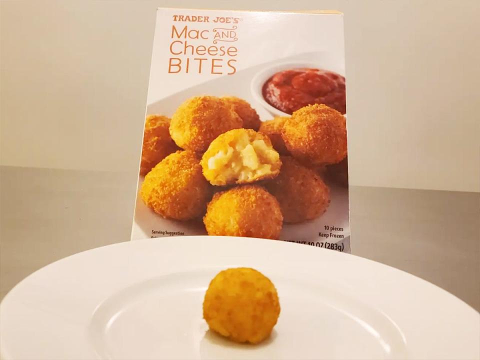 A golden-brown mac-and-cheese bite ball sits on a white plate in front of a gray box of mac and cheese bites