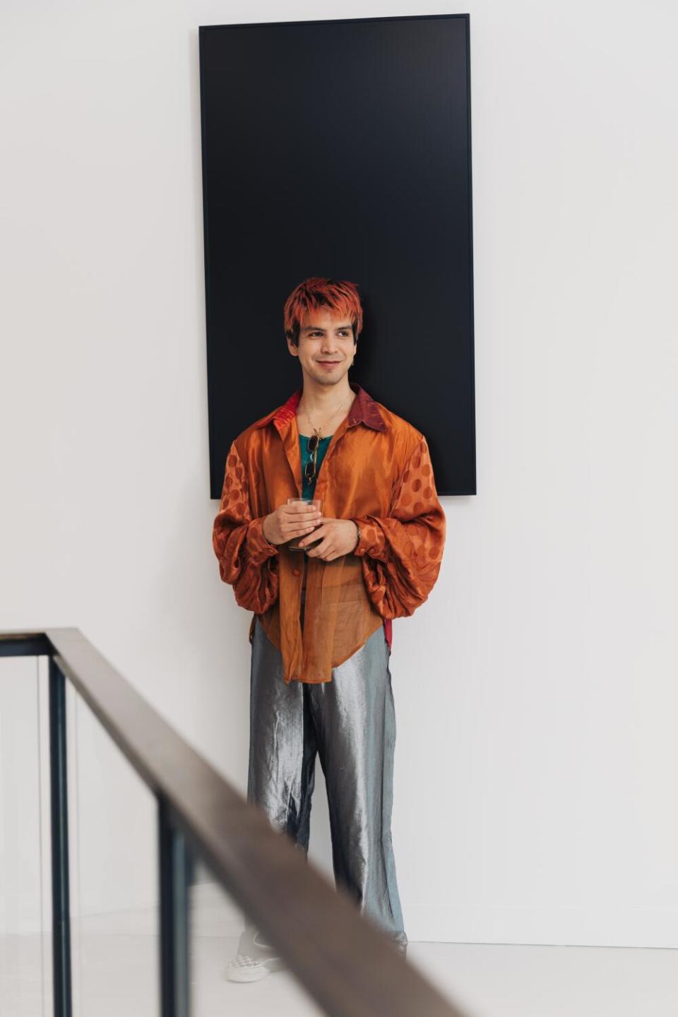 Julio Torres, in silky grey pants and transparent orange shirt, stands before a black painting in a white room.