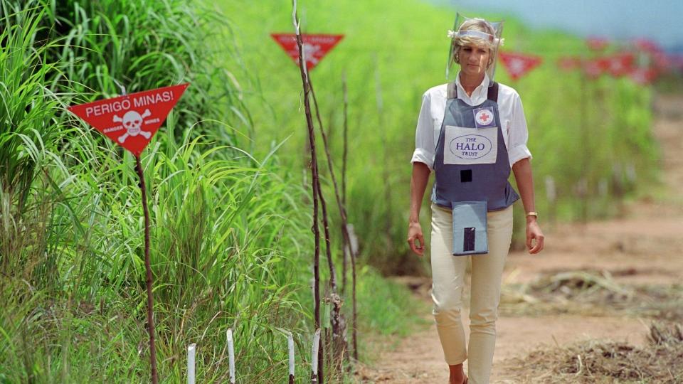 Princes Diana in angola walking past a field of mines, she's wearing protective gear and the grass is tall next to her