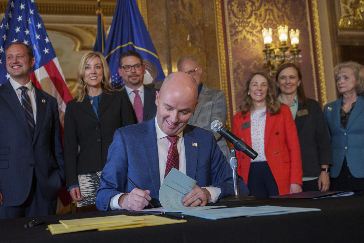 Gov. Spencer Cox, surrounded by a group of people looking upbeat, enthusiastically signs a document in a gilded hall.