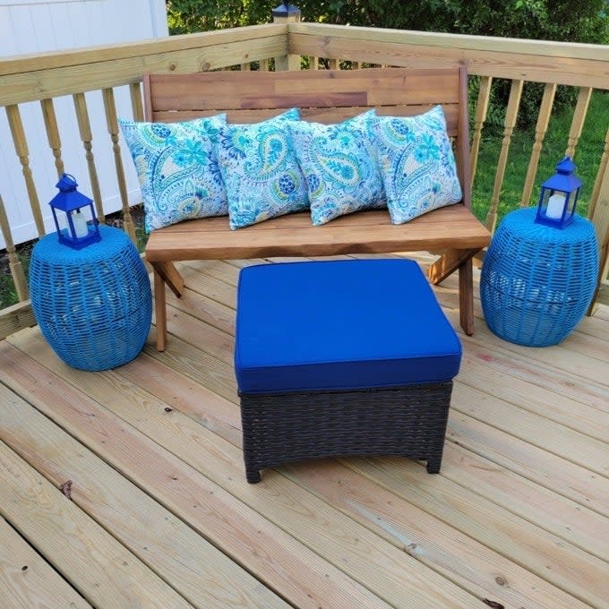 Outdoor furniture setup with a wooden bench, blue cushions, two lanterns, and a wicker ottoman