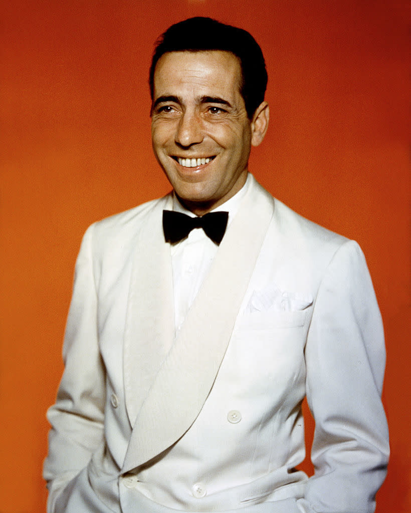 A smiling man in a white tuxedo jacket with a black bow tie against an orange background