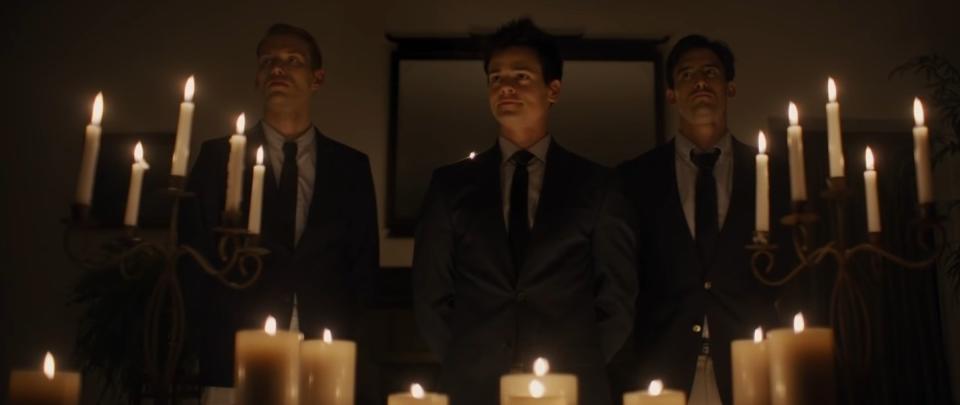 Three scary-looking fraternity brothers stand in full suits in front of a candlelit room in "Pledge"