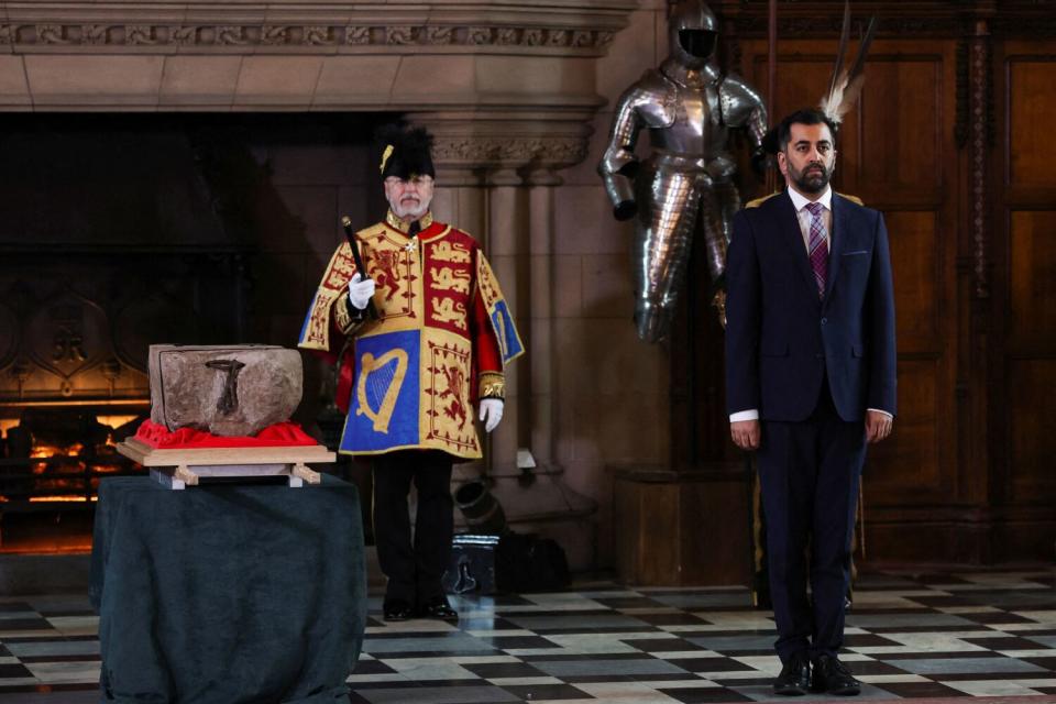 A man with a dark beard, in a dark suit and tie, stands next to a stone on a stand, guarded by a man in colorful uniform