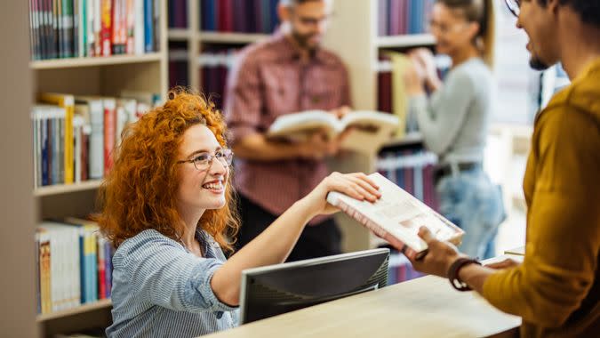 Happy female librarian taking a book from a male student in the library.