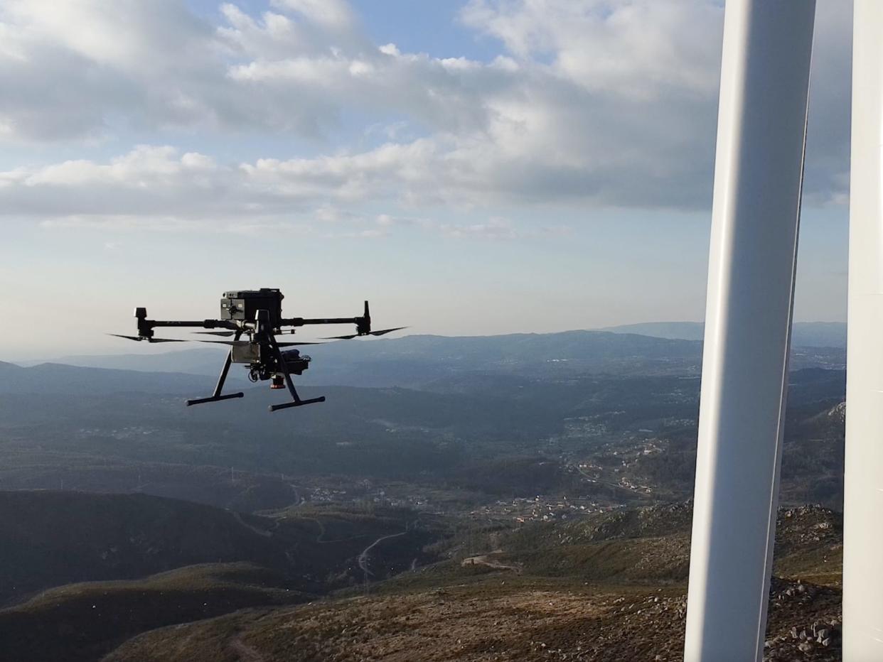 A black drone flies near a white wind turbine in the mountains.