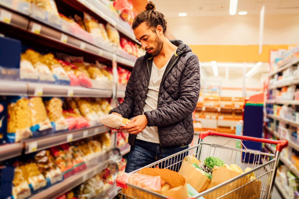 Young bearded man in groceries shopping. He is choosing pasta and reading nutrition label on product. Location released.