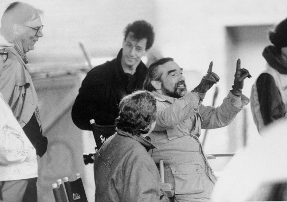 a black and white photo of a bearded martin scorsese pointing upwards and smiling as he directs a film, with several people around him