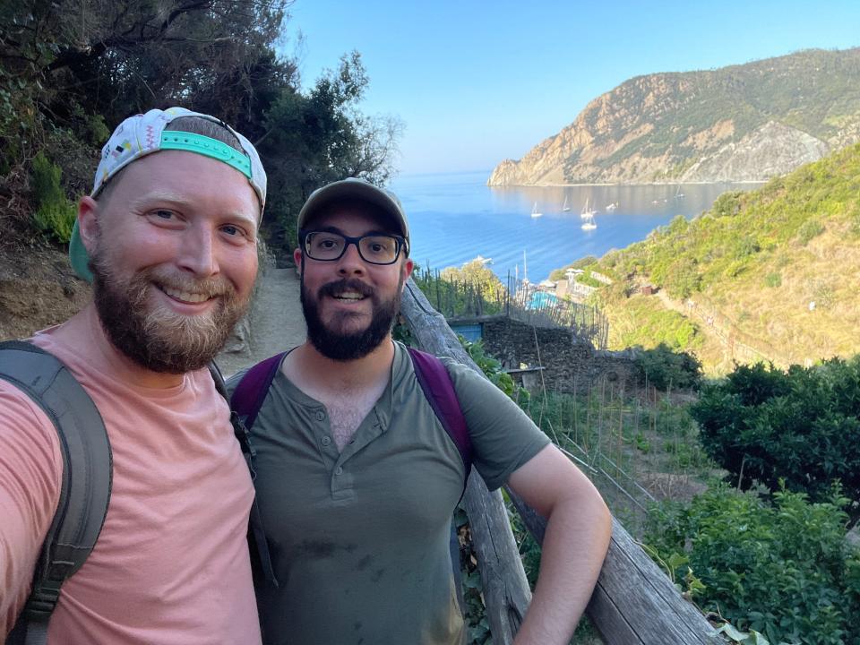 The author Timothy Moore and his husband smiling at the top of a Cinque Terre hike with blue waters and green hills in the background