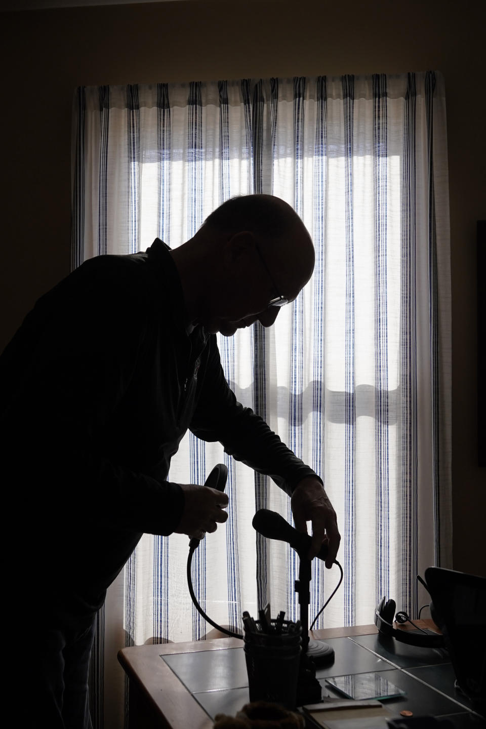 Roger Strukhoff 67, stands in silhouette cleaning off his home office desk Thursday, Jan. 20, 2022, in his DeKalb, Ill., home. Strukhoff was being treated for intestinal bleeding at a hospital outside Chicago this month when he suffered a mild heart attack. Normally, the medical staff would have sent Strukhoff to the intensive care unit, but, overrun with COVID-19 patients, the staff instead had to wheel a heart monitor into his room and quickly administer nitroglycerin and morphine. (AP Photo/Charles Rex Arbogast)