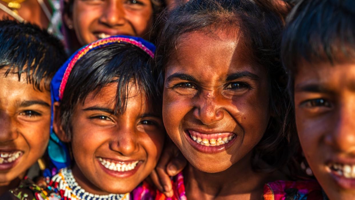  Close-up image of a group of four Indian children (with the face of another more blurred behind them) who are smiling at the camera. The sun in shining on their faces. 