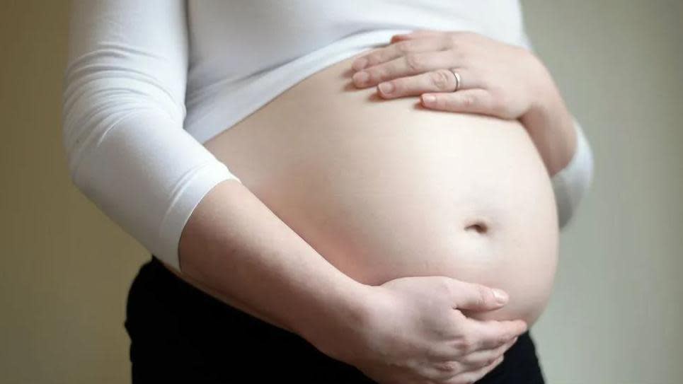 A woman holds her pregnant stomach. She wears a white top, black trousers and a wedding ring on her left hand.