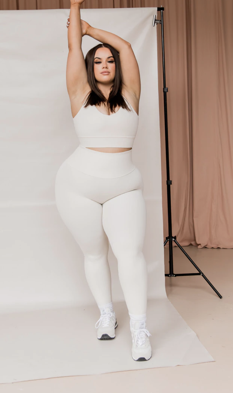 A model wears a white pair of Leo Leggings, $79 and a matching crop top as she faces the camera with hands overhead.