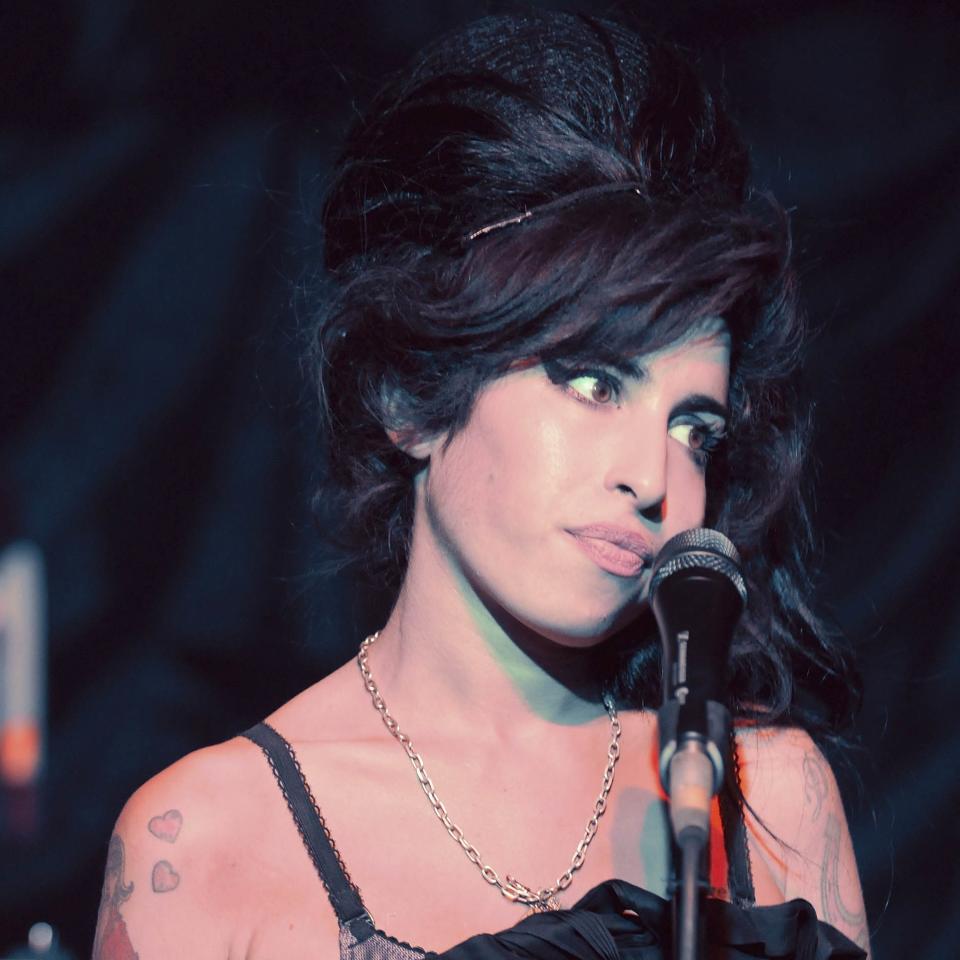 Amy Winehouse with beehive hairstyle, standing by a microphone, expression pensive