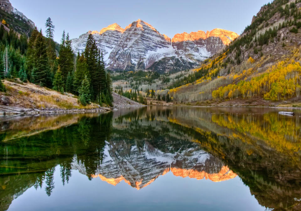 Sunrise hits snow dusted peaks of Maroon Bells while being reflected in lake below, along with Aspen trees in their full fall foliage display of golden colors.