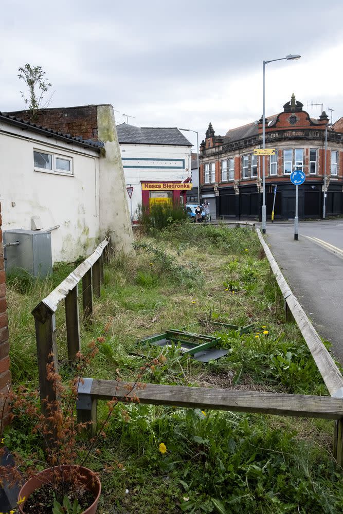 a fenced in area of urban scrubland next to a road with commercial buildings in the background