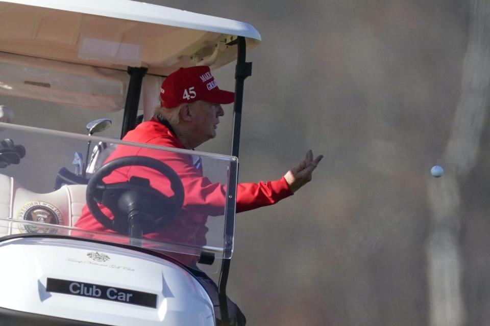 Donald Trump, in a red jacket wearing a red Make America Great baseball cap, tosses a golf ball from a golf cart.