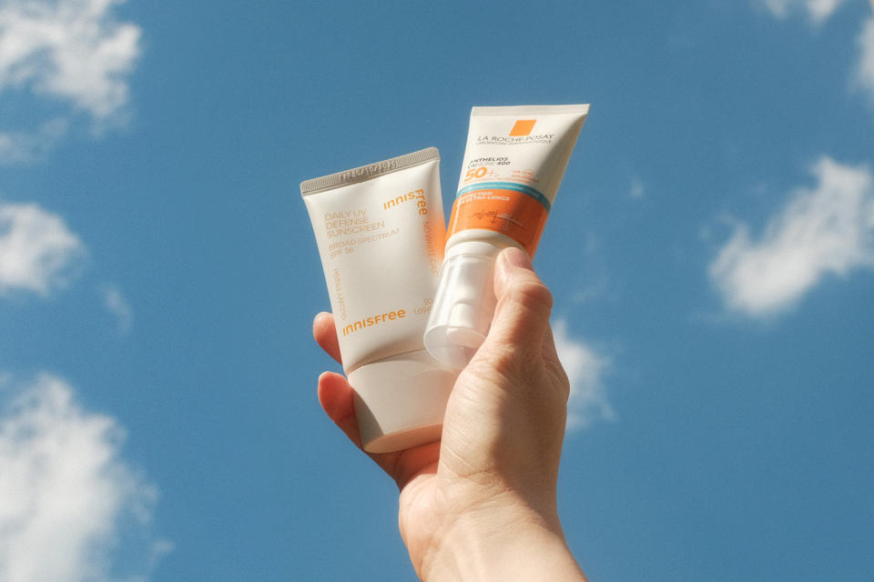 A hand holds two bottles of sunscreen against a blue sky with clouds (Chelsea Stahl and Elise Wrabetz / NBC News)