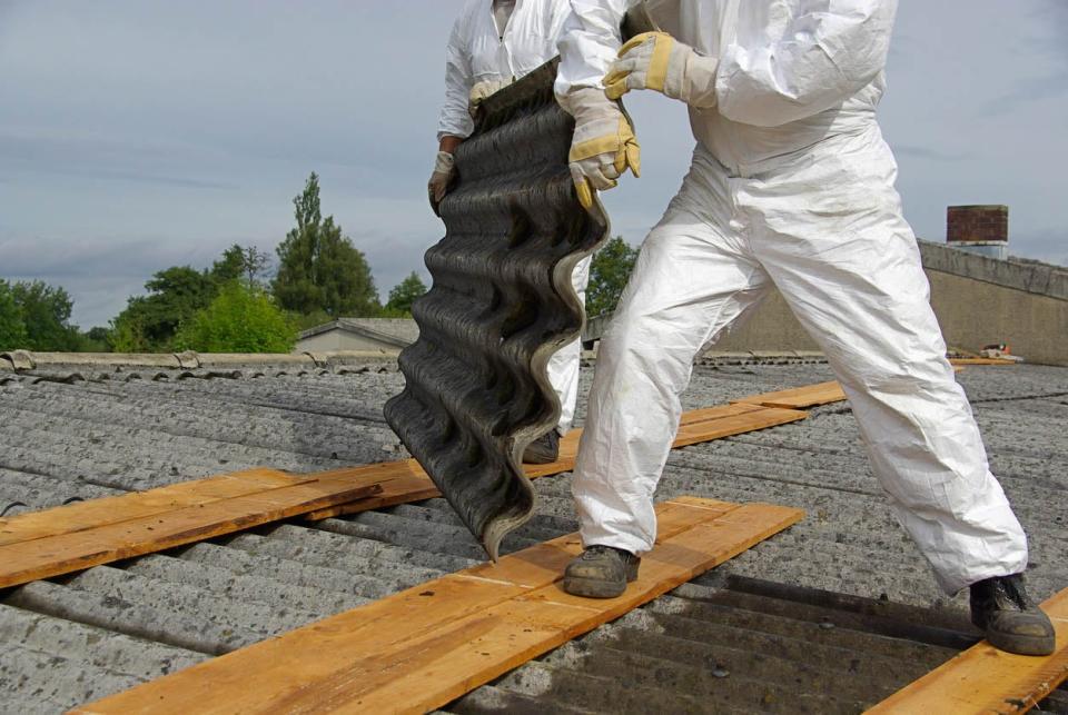 A worker in a white jumpsuit removes asbestos materials form a roof.